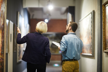 Porträtansicht von hinten: Zwei Museumsmitarbeiter, die sich über Gemälde unterhalten, während sie durch eine Kunstgalerie gehen